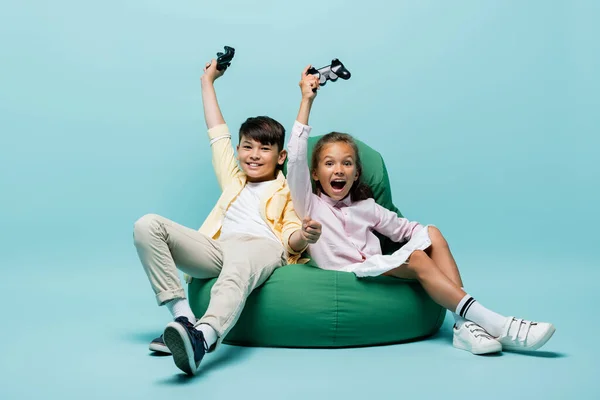 KYIV, UCRANIA - 2 de julio de 2021: Niños interracial excitados sosteniendo joysticks en una silla de frijoles sobre fondo azul - foto de stock
