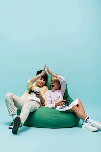 KYIV, UCRANIA - 2 DE JULIO DE 2021: Niños multiétnicos sosteniendo joysticks y dando cinco altos en la silla del bolso de frijoles sobre fondo azul — Stock Photo