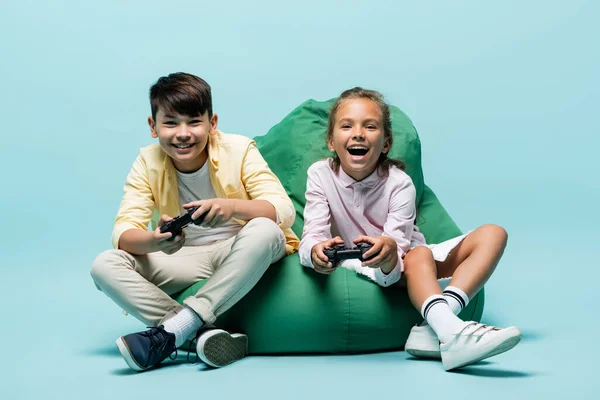 KYIV, UCRANIA - 2 de julio de 2021: Felices niños preadolescentes multiétnicos jugando videojuegos en una silla de frijoles sobre fondo azul - foto de stock