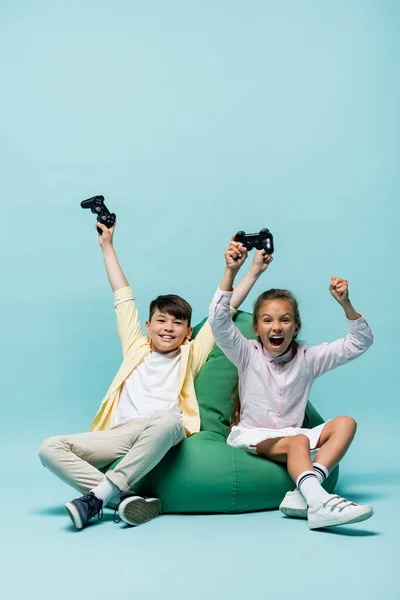 KYIV, UCRANIA - 2 DE JULIO DE 2021: Niños multiétnicos emocionados sosteniendo joysticks en la silla del bolso de frijoles sobre fondo azul - foto de stock