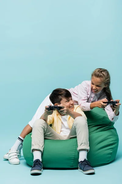 KYIV, UCRANIA - 2 de julio de 2021: Un niño asiático sonriente jugando un videojuego con un amigo en una silla de frijoles sobre fondo azul - foto de stock