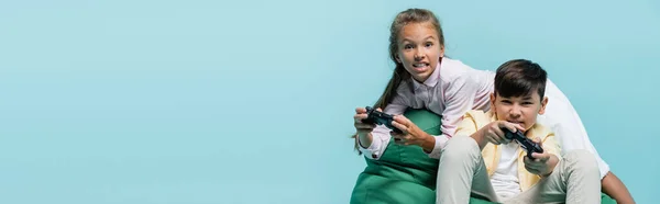 КИЕВ, УКРАИНА - 2 ИЮЛЯ 2021 года: Концентрированный ребенок играет в видеоигру с азиатским другом изолированы на синий, баннер — стоковое фото