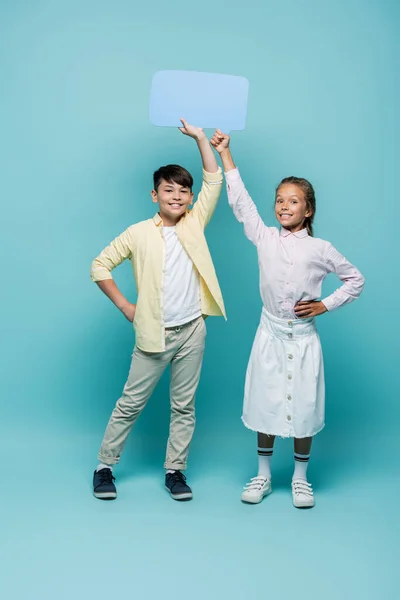 Smiling multiethnic schoolkids holding speech bubble on blue background — Stock Photo