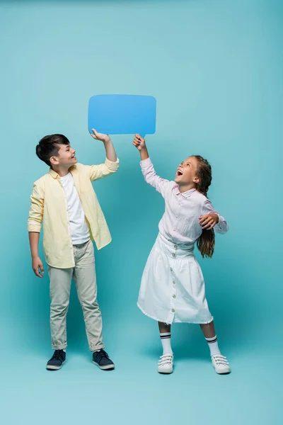Веселые межрасовые школьники держат пузырь речи на синем фоне — стоковое фото