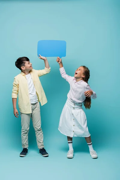 Alegre interracial colegiales celebración discurso burbuja en azul fondo - foto de stock