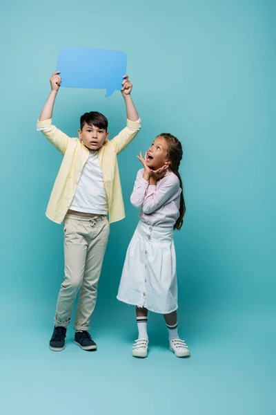 Shocked asian schoolboy holding speech bubble near friend on blue background — Stock Photo