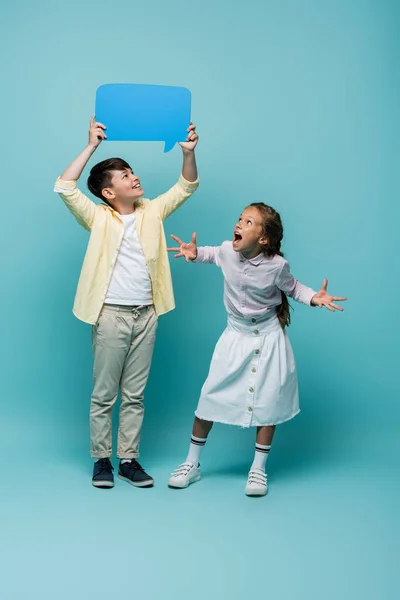 Excited and smiling interracial pupils looking at speech bubble on blue background — Stock Photo