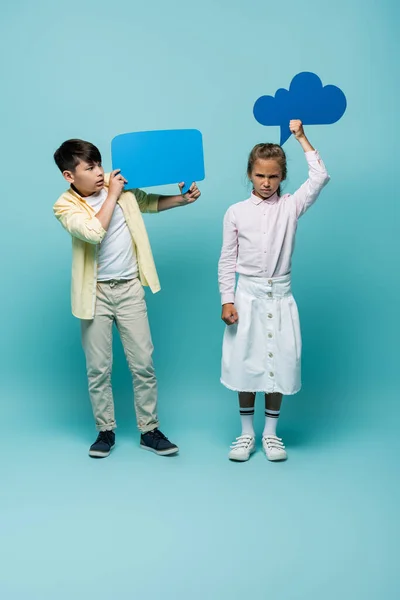 Enojado colegiala celebración pensamiento burbuja cerca asiático amigo en azul fondo - foto de stock