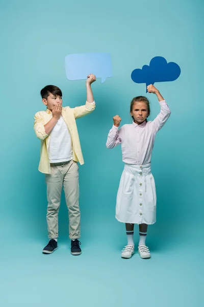 Aggressive schoolgirl holding thought bubble near scared asian friend on blue background — Stock Photo