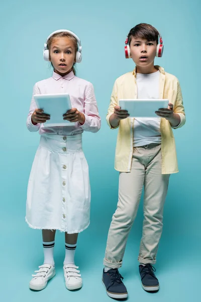 Schockierte Schüler mit Kopfhörern halten digitale Tablets auf blauem Grund — Stockfoto