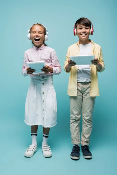 Positive multiethnische Schüler mit Kopfhörern, die digitale Tablets auf blau halten — Stockfoto