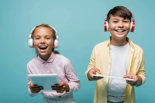 Alunos inter-raciais felizes em fones de ouvido segurando tablets digitais isolados em azul — Fotografia de Stock