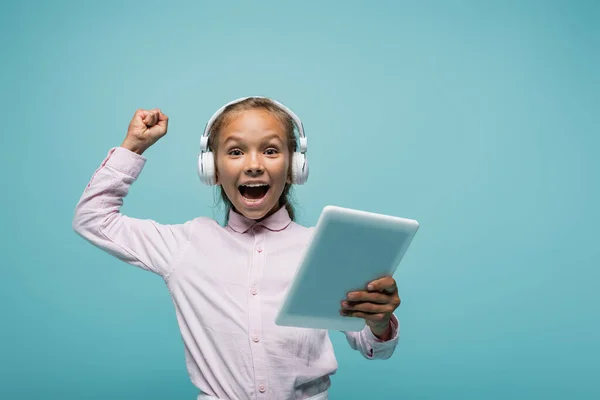 Aufgeregte Schülerin mit Kopfhörer hält digitales Tablet isoliert auf blauem Grund — Stockfoto