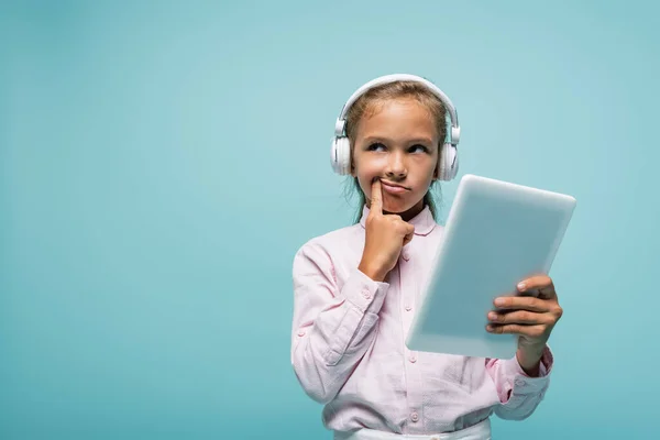 Pensive schoolkid in headphones holding digital tablet isolated on blue — Stock Photo