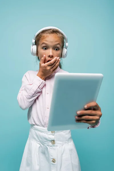 Schockiertes Schulmädchen mit Kopfhörern blickt auf digitales Tablet isoliert auf blauem Grund — Stockfoto