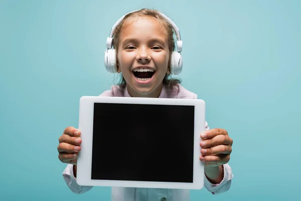 Lächelndes Schulmädchen mit Kopfhörern blickt in die Kamera und hält ein digitales Tablet isoliert auf blauem Grund — Stockfoto