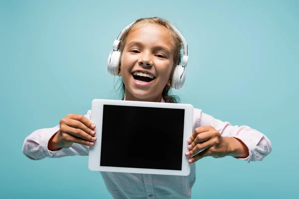 Positive preteen schoolgirl in headphones holding digital tablet isolated on blue — Stock Photo