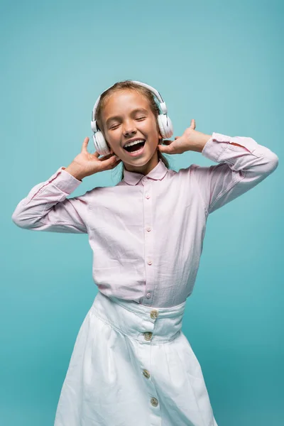 Colegiala positiva escuchando música en auriculares aislados en azul - foto de stock