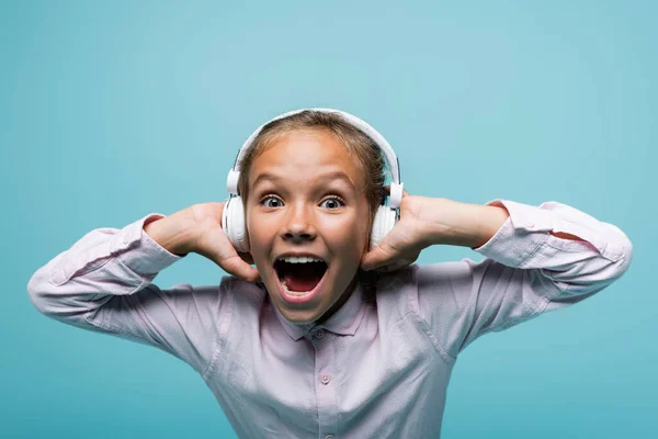 Emocionado preadolescente escolar en los auriculares mirando a la cámara aislada en azul - foto de stock