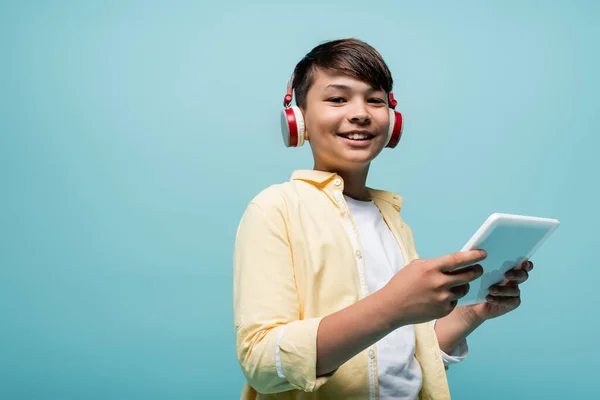 Sourire asiatique écolier dans casque tenant numérique tablette isolé sur bleu — Photo de stock