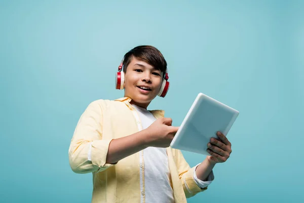 Low angle view of smiling asian schoolboy in headphones holding digital tablet isolated on blue — Stock Photo