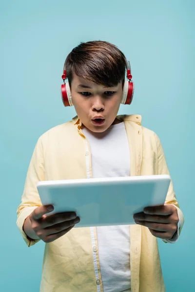 Shocked asian schoolkid in headphones using digital tablet isolated on blue — Stock Photo