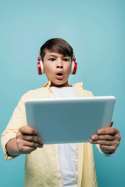 Tiefansicht eines schockierten asiatischen Schülers in Kopfhörern, der verschwommenes digitales Tablet isoliert auf blauem Grund hält — Stockfoto