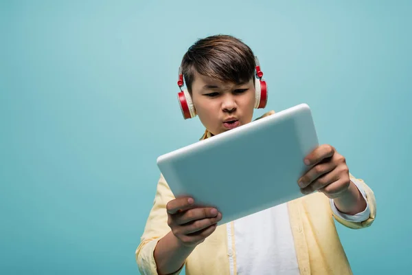 Enfocado asiático escolar en auriculares usando digital tablet aislado en azul - foto de stock