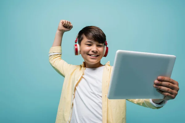 Positive asian schoolboy in headphones showing yes gesture and holding digital tablet isolated on blue — Stock Photo