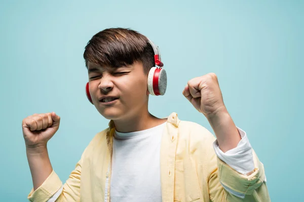 Aufgeregtes asiatisches Kind hört Musik über Kopfhörer und zeigt Ja-Geste isoliert auf blauem Grund — Stockfoto