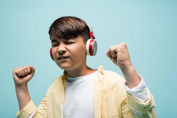 Excited asian child listening music in headphones and showing yes gesture isolated on blue — Stock Photo