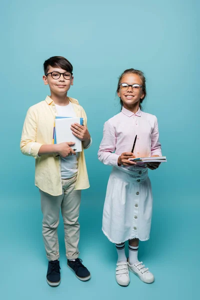 Lächelnde interrassische Schulkinder mit Notizbüchern in der Hand und Blick in die Kamera auf blauem Hintergrund — Stockfoto