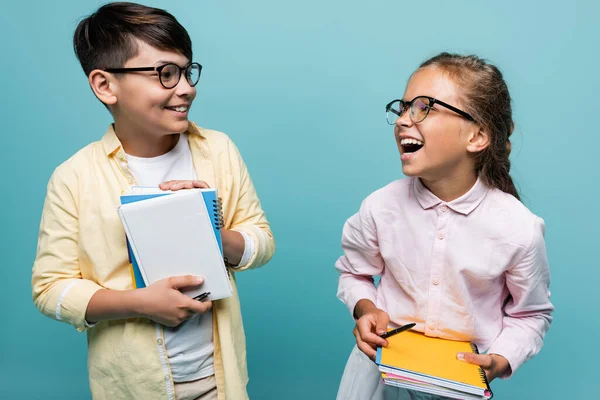 Positive multiethnic schoolkids in eyeglasses holding notebooks isolated on blue — Stock Photo