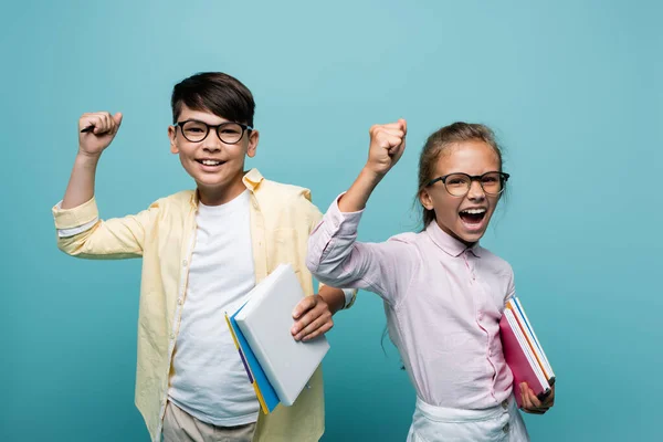 Eccitato interrazziale scolari in occhiali che tengono quaderni e mostrando sì gesto isolato su blu — Stock Photo