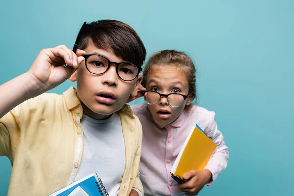 Konzentrierte interrassische Schulkinder in Brillen, Notizbücher in der Hand und Kamera isoliert auf blauem Grund betrachtend — Stockfoto