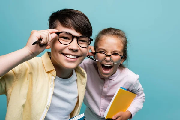 Веселі багатоетнічні школярі в окулярах тримають ручки і блокноти ізольовані на синьому — стокове фото