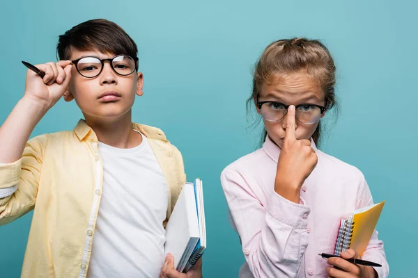 Interrassische Schüler justieren Brillen und halten Hefte isoliert auf blauem Grund — Stockfoto