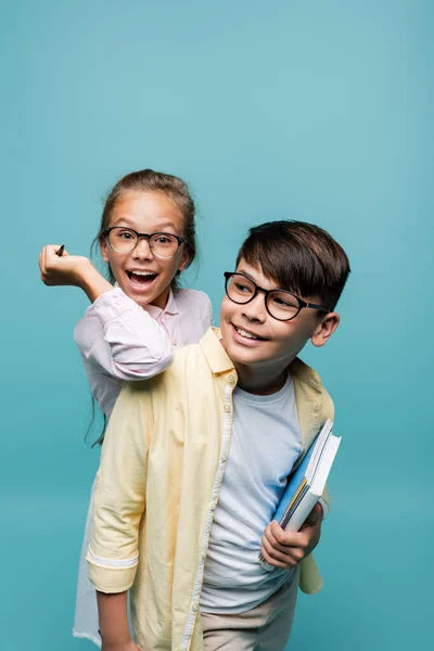 Cheerful schoolgirl in eyeglasses holding pen near asian classmate with notebooks isolated on blue — Stock Photo