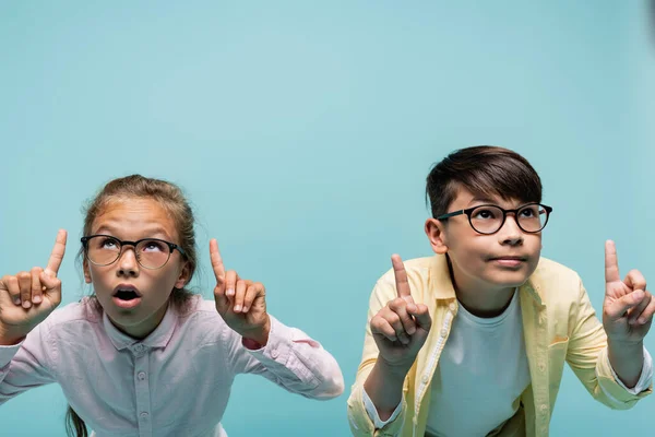 Interracial schoolkids in eyeglasses pointing with fingers isolated on blue — Stock Photo