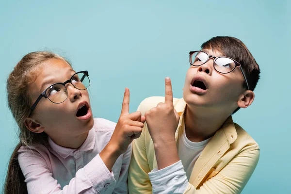 Shocked interracial schoolkids in eyeglasses pointing with fingers isolated on blue — Stock Photo