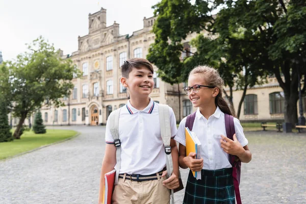Escolares multiétnicos sonrientes con cuadernos caminando al aire libre - foto de stock