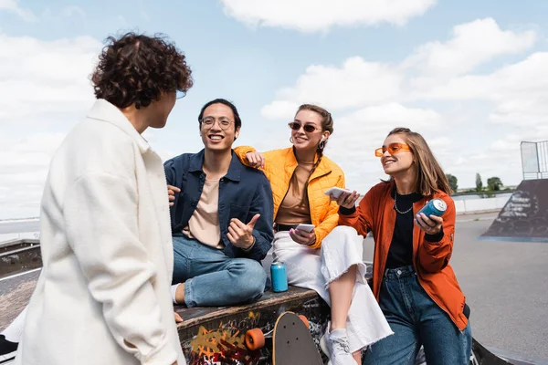 Sonriente asiático hombre mostrando pulgar arriba cerca de amigos con smartphones y soda latas - foto de stock