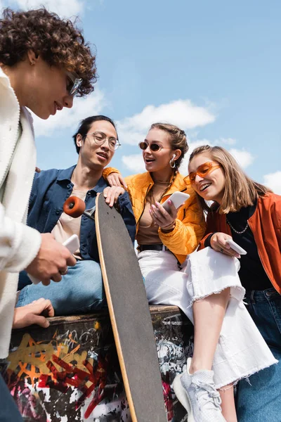 Young asian man with longboard sitting with cheerful and stylish friends outdoors — Stock Photo