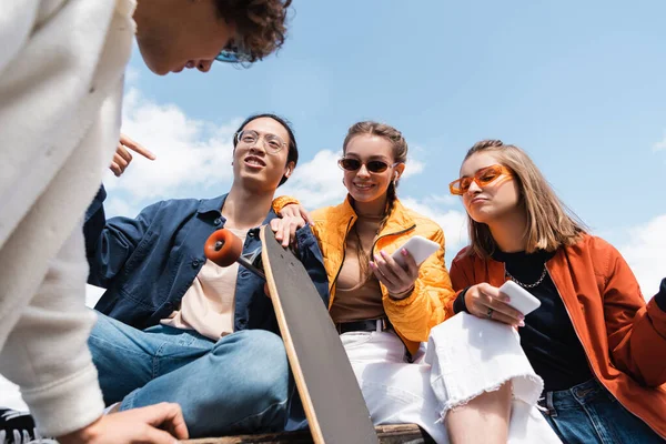 Low angle view of asian skater pointing with finger near friends with smartphones outdoors — Stock Photo