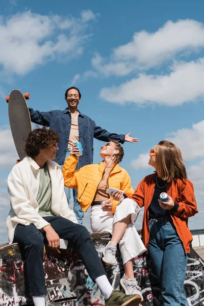 Asian man with longboard standing with outstretched hands above happy friends with smartphones and soda cans — Stock Photo