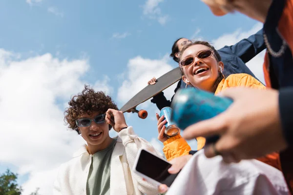 Vista a basso angolo di gioiosi amici multietnici con lattina di soda e smartphone all'aperto — Foto stock