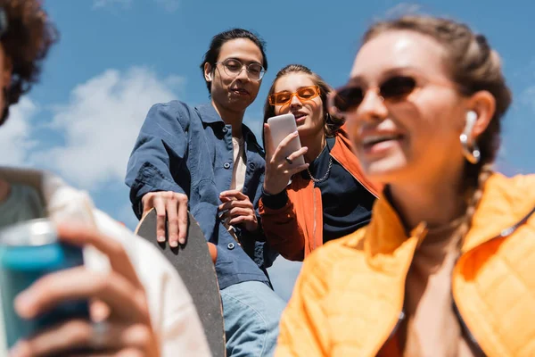 Low angle view of young woman taking selfie with asian skater near friends on blurred foreground — Stock Photo