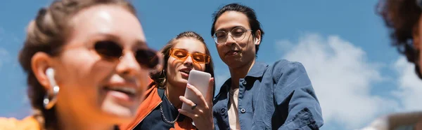 Junge Frau und asiatischer Mann schauen auf Smartphone in der Nähe verschwommener Freunde, Banner — Stockfoto