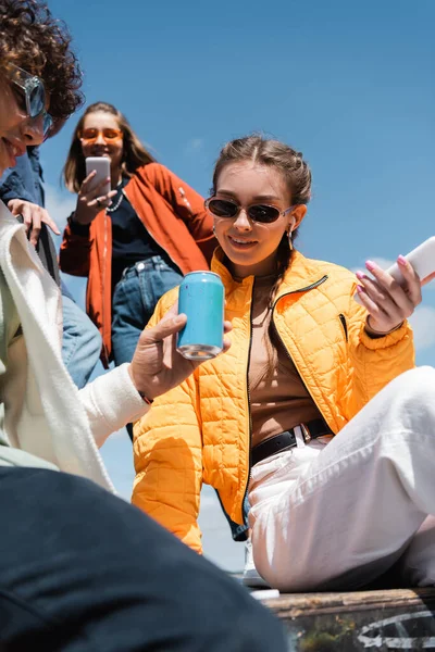 Vista de ángulo bajo de amigos jóvenes y de moda con gadgets y latas de refrescos contra el cielo azul - foto de stock