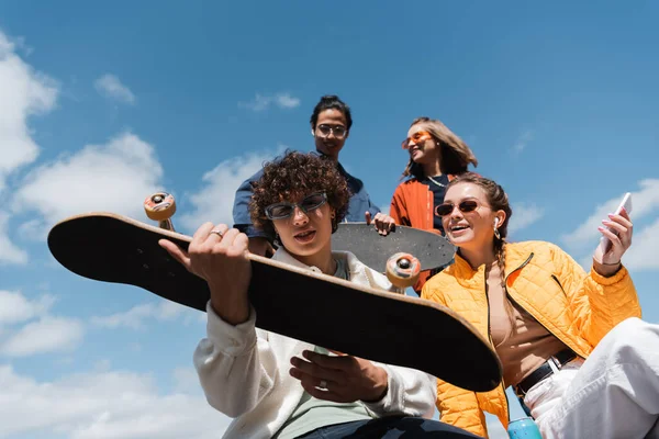 Low angle view of young man holding skateboard near happy interracial friends outdoors — Stock Photo